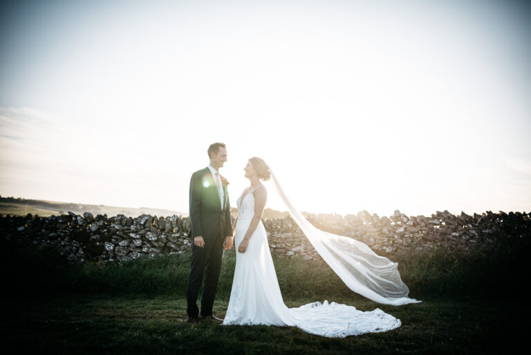couple standing by a dry stone wall in a field at golden hour, brides veil flowing in the breeze