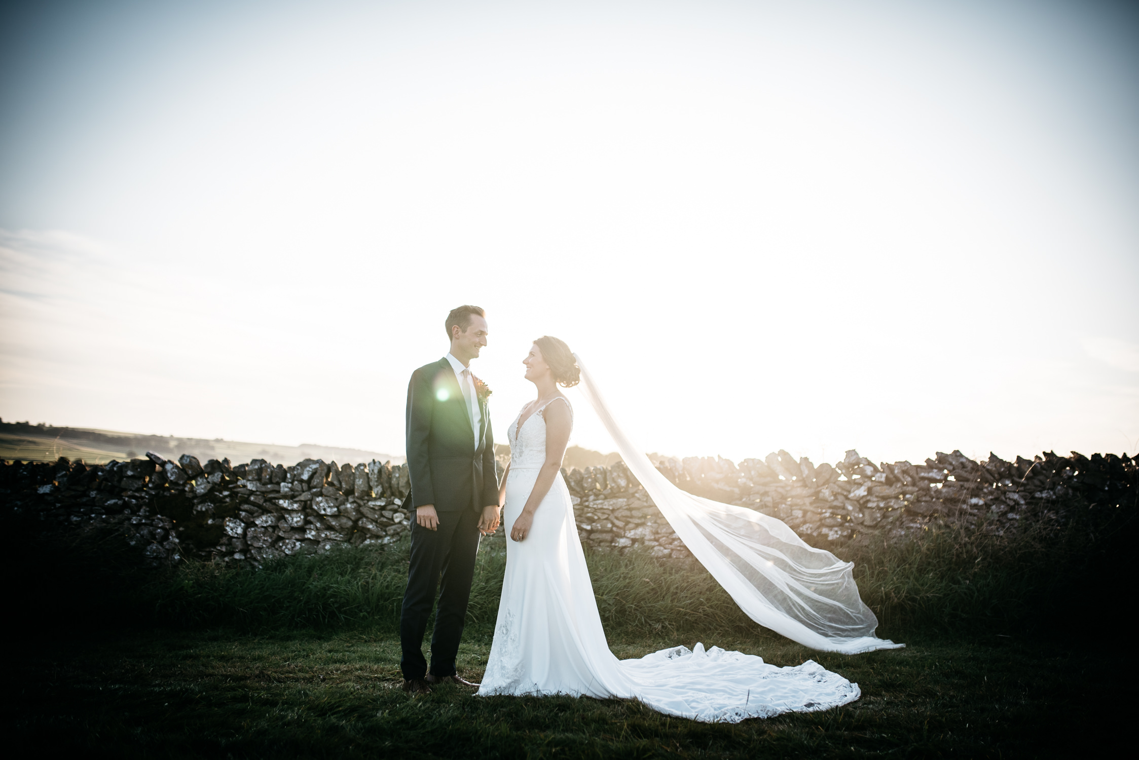 couple standing by a dry stone wall in a field at golden hour, brides veil flowing in the breeze