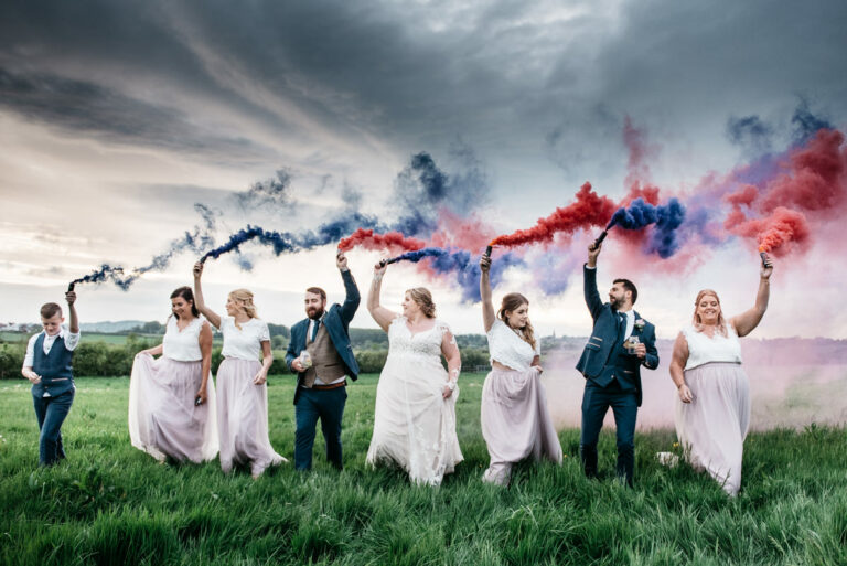 Cool bridal party walking in a field holding colourful smoke bombs aloft