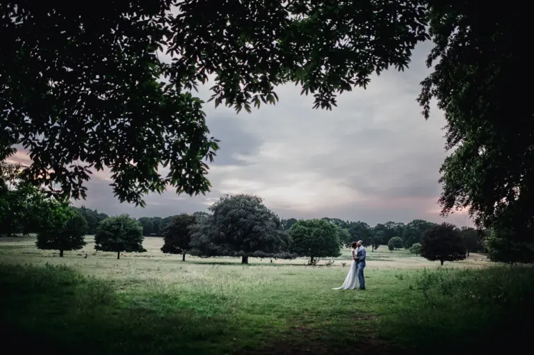 Wide landscape shot of a Bride and Groom kiss on Wollaton Park with sunset and deer behind